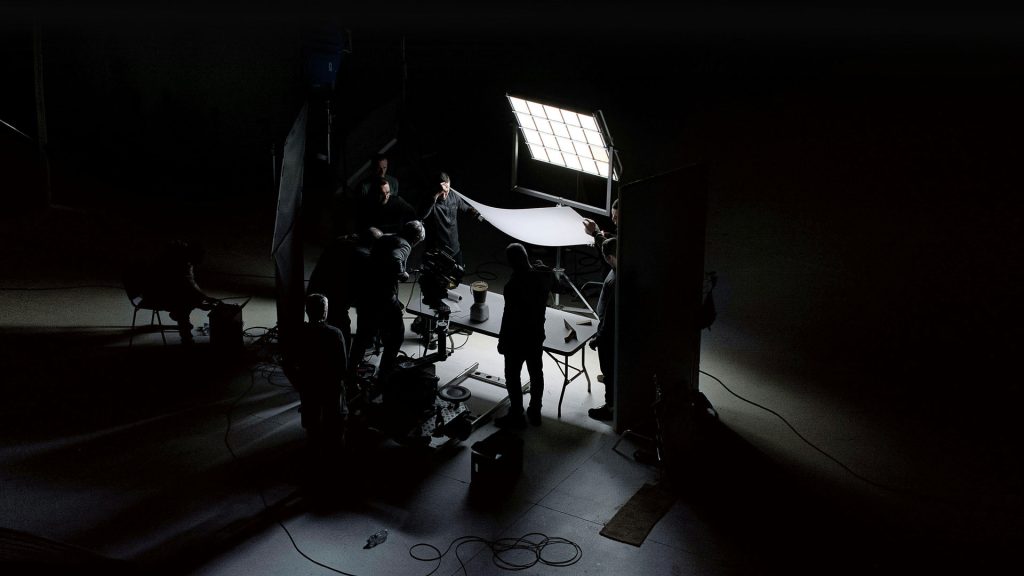 Hier siehst du das Team von Background-Photodesign bei der Arbeit im Studio in Leipzig, wo sorgfältig mit professionellem Beleuchtungs- und Kameraequipment hantiert wird. Die konzentrierte Stimmung wird von der akkuraten Anordnung des LED-Panels und des Diffusors unterstrichen, während sich Kabel geordnet über den Studioboden ziehen und Schatten an den Wänden kreatives Leben einhauchen. Produktionsset Beleuchtungsequipment LED-Panel Diffusor Kameraausrüstung Stativ Crewmitglieder Dunkler Raum Kabel Arbeitstische Reflexionsschirm Techniker Filmproduktion Fotostudio Schatten und Lichtspiel Hinter den Kulissen Konzentration Teamarbeit Medienproduktion Kreative Industrie
